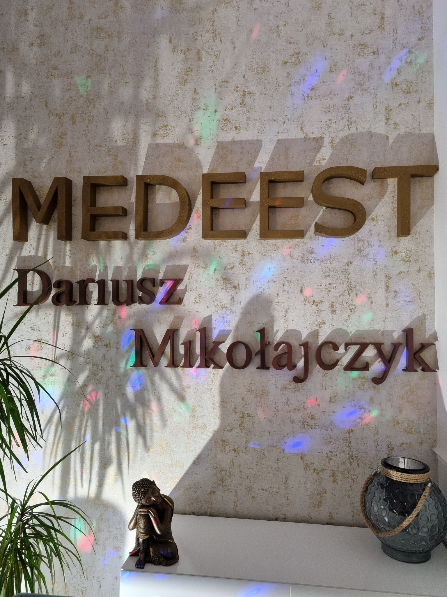 Ściana z dekoracyjnymi, przestrzennymi napisami MEDEEST Dariusz Mikołajczyk, oświetlona kolorowymi refleksami świetlnymi, z rośliną doniczkową i figurką Buddy na białej komodzie.