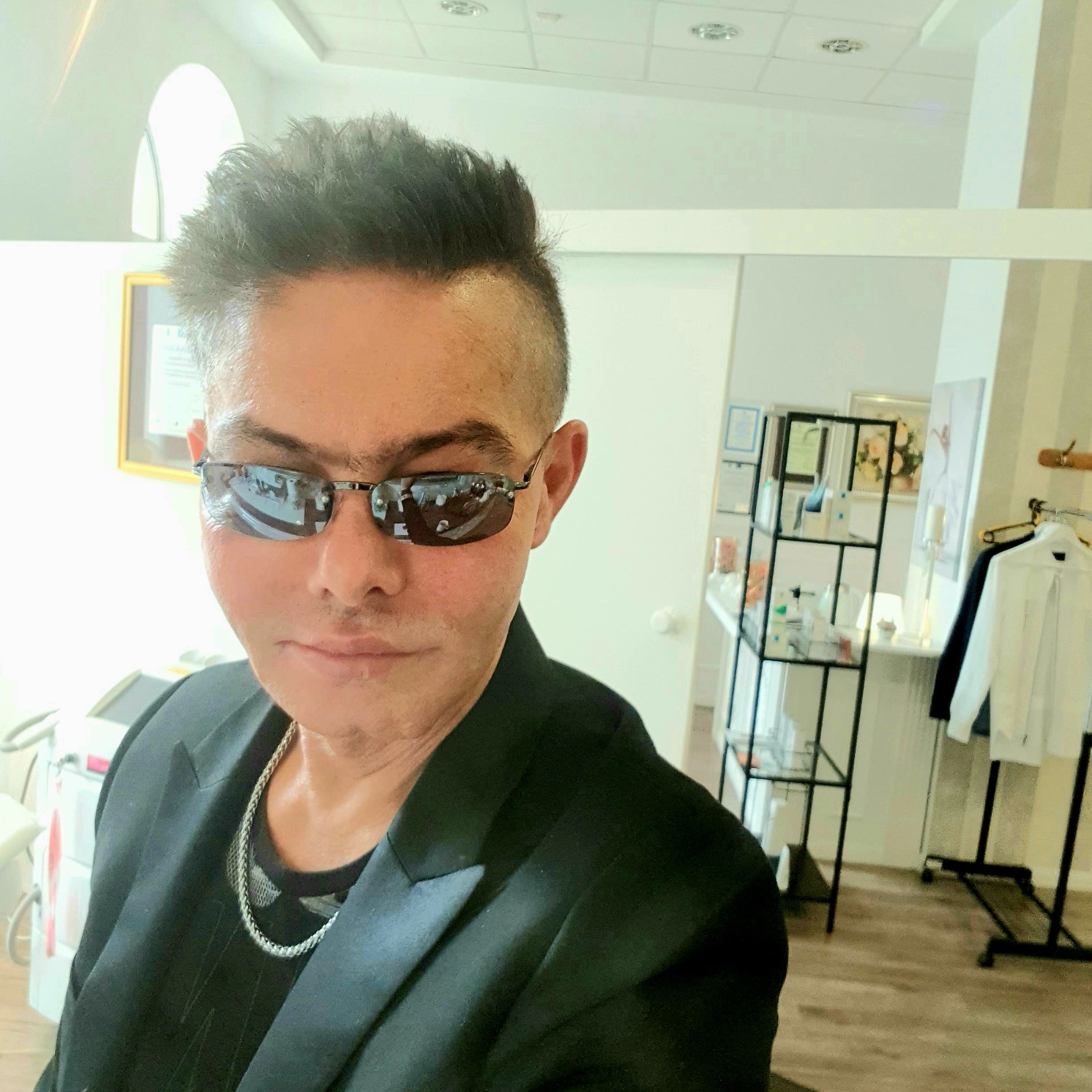 Mężczyzna w okularach przeciwsłonecznych i czarnej marynarce, w tle gabinet z urządzeniami medycznymi, możliwe że po zabiegu medycyny estetycznej.