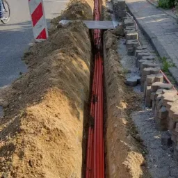 Długi, wąski wykop wzdłuż ulicy, z widocznymi pomarańczowymi rurami ułożonymi na dnie, zabezpieczony z jednej strony kostką brukową i znakami ostrzegawczymi.