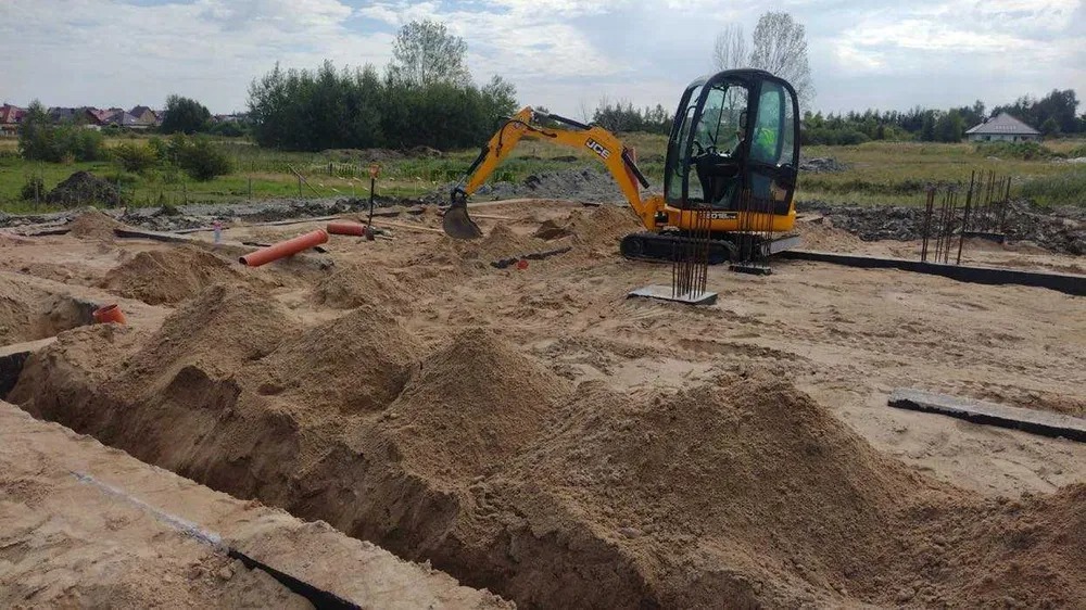 Żółta mini koparka JCB w trakcie prac ziemnych na placu budowy, widoczne wykopane rowy, wystające pręty zbrojeniowe i rury kanalizacyjne.