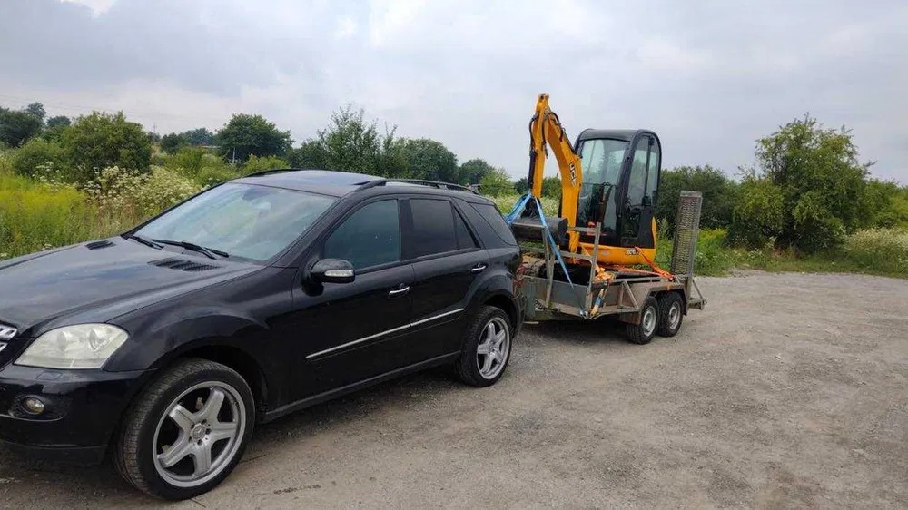 Czarny SUV Mercedes ciągnie na przyczepie żółtą mini koparkę JCB, transport sprzętu budowlanego na tle zieleni.