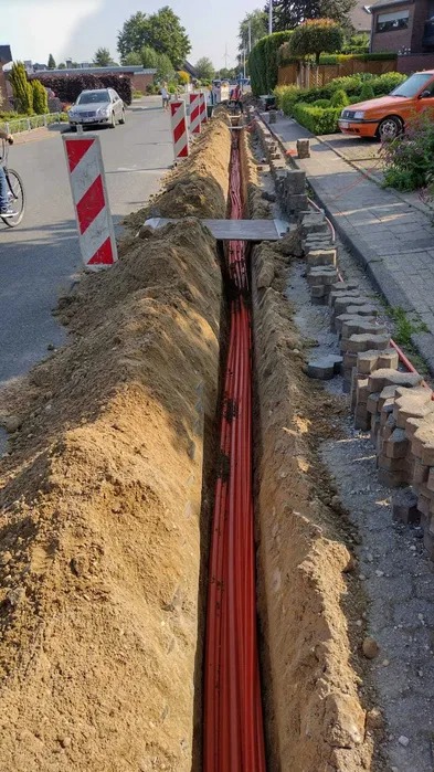 Długi, wąski wykop wzdłuż ulicy, z widocznymi pomarańczowymi rurami ułożonymi na dnie, zabezpieczony z jednej strony kostką brukową i znakami ostrzegawczymi.