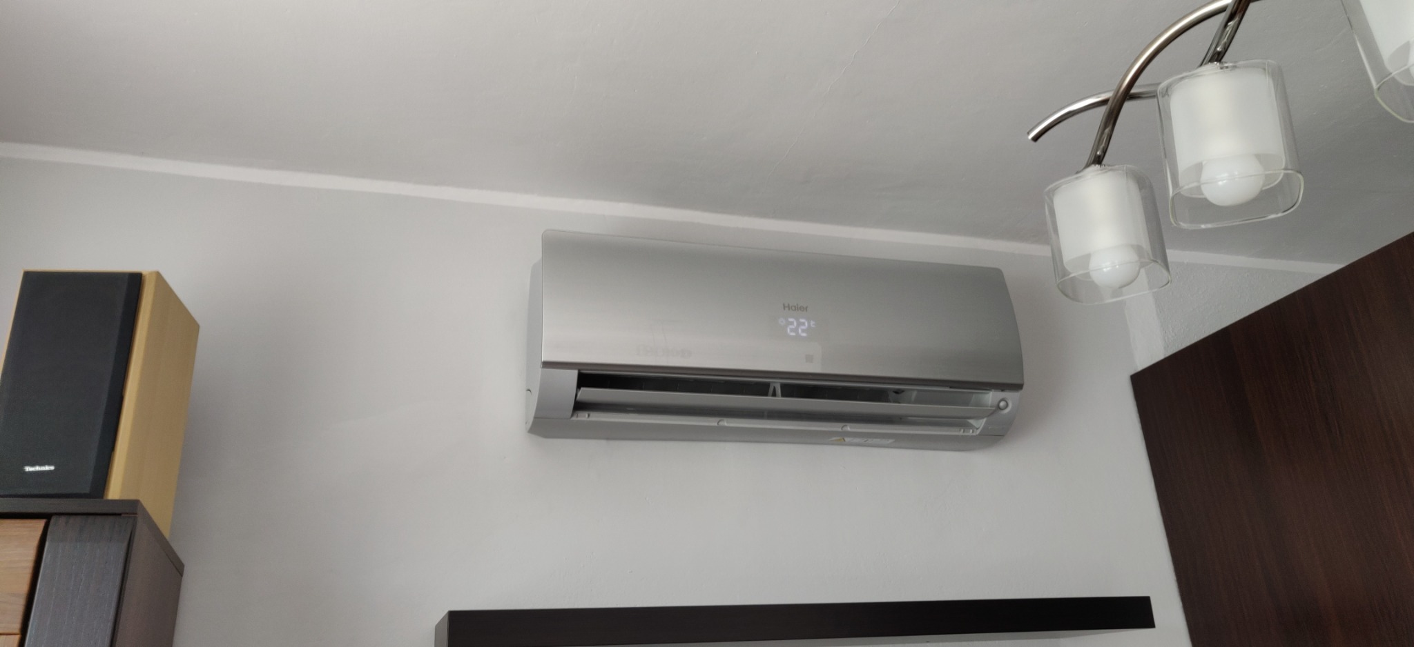 Zamontowany klimatyzator Haier w szarym kolorze na białej ścianie, z widocznym wyświetlaczem temperatury 22 stopnie, obok widoczny głośnik Technics i fragment lampy sufitowej.