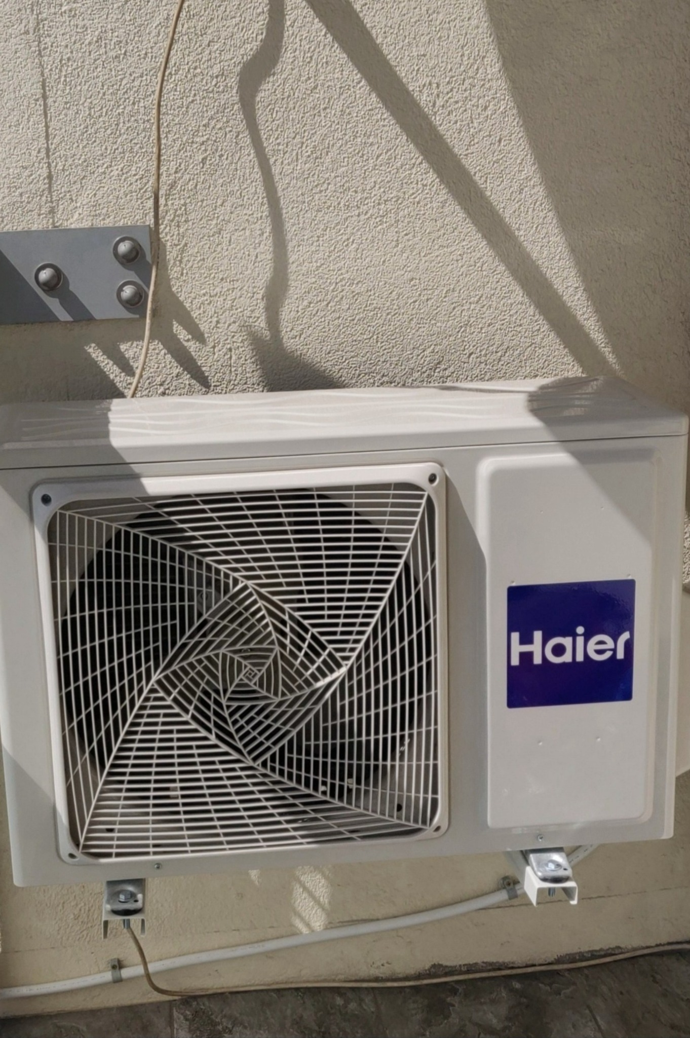 Biała jednostka zewnętrzna klimatyzatora Haier zamontowana na ścianie budynku, widoczne przewody i cień na elewacji.