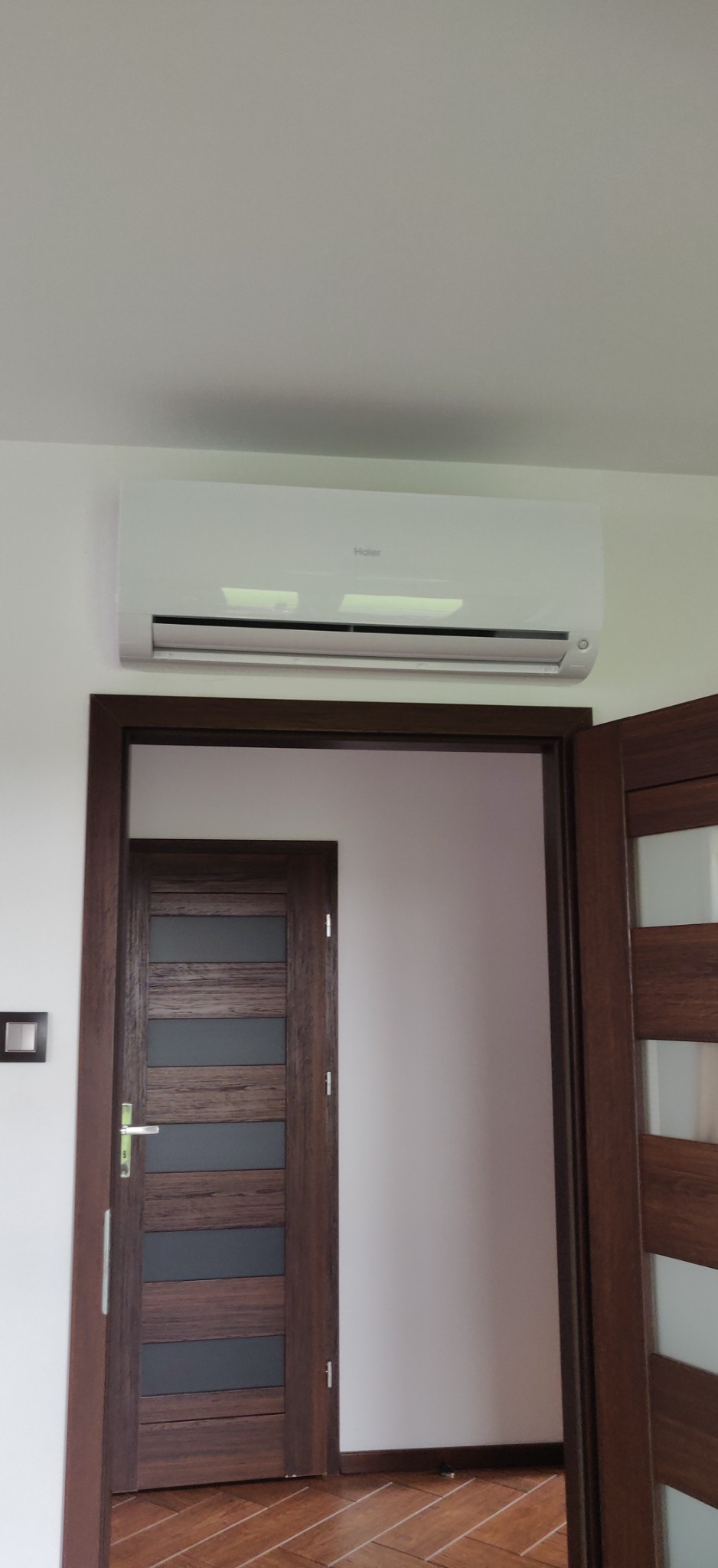 Biały klimatyzator Haier zamontowany nad futryną drzwiową w jasnym wnętrzu, widoczna druga para drzwi w tle.