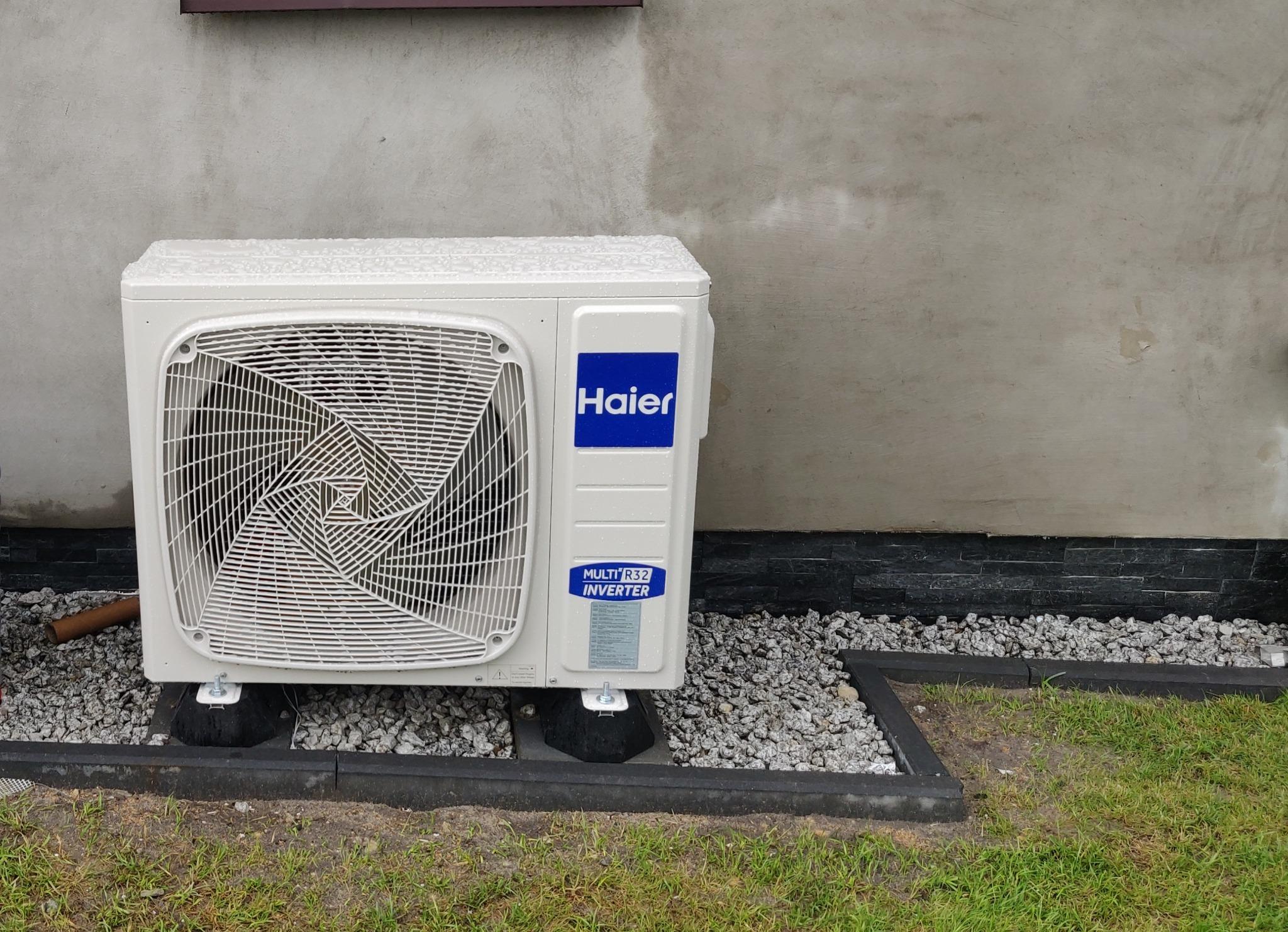 Zewnętrzna jednostka klimatyzacji Haier Multi R32 Inverter, ustawiona na gumowych podstawach na żwirowym podłożu przy ścianie budynku z czarną kamienną okładziną.