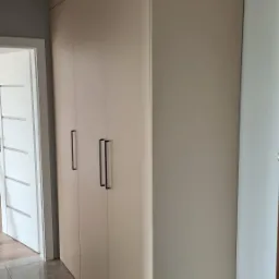 Beżowa szafa wnękowa z trzema drzwiami i czarnymi, podłużnymi uchwytami na tle szarej podłogi i białych drzwi.