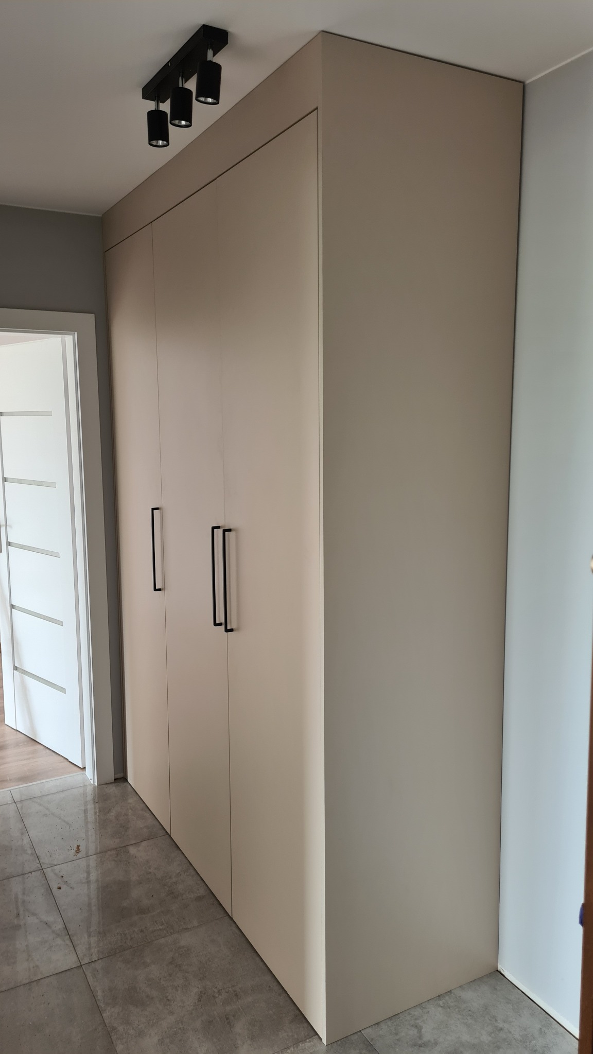 Beżowa szafa wnękowa z trzema drzwiami i czarnymi, podłużnymi uchwytami na tle szarej podłogi i białych drzwi.