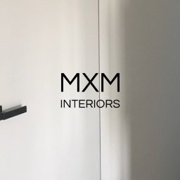Mxm Interiors - Białe, minimalistyczne drzwi z czarnymi klamkami i logo MXM Interiors, częściowo otwarte, ukazujące szare wnętrze pomieszczenia.
