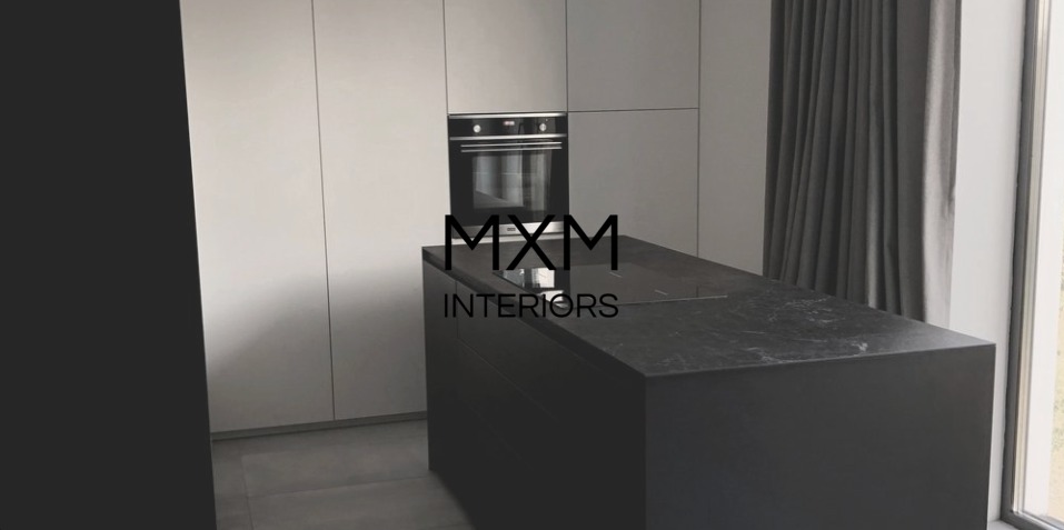 Minimalistyczna kuchnia z czarnym marmurowym blatem wyspy, wbudowanym piekarnikiem i białymi szafkami, oświetlona naturalnym światłem z okna z szarą zasłoną.