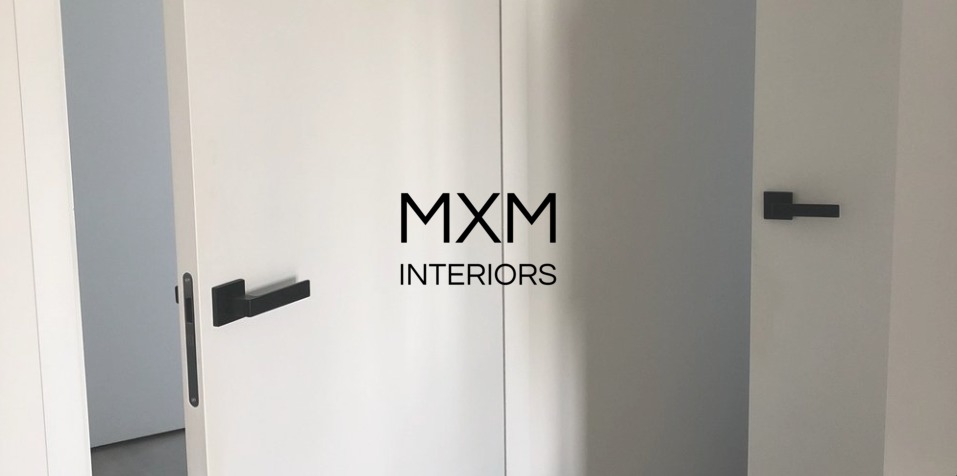 Białe, minimalistyczne drzwi z czarnymi klamkami i logo MXM Interiors, częściowo otwarte, ukazujące szare wnętrze pomieszczenia.