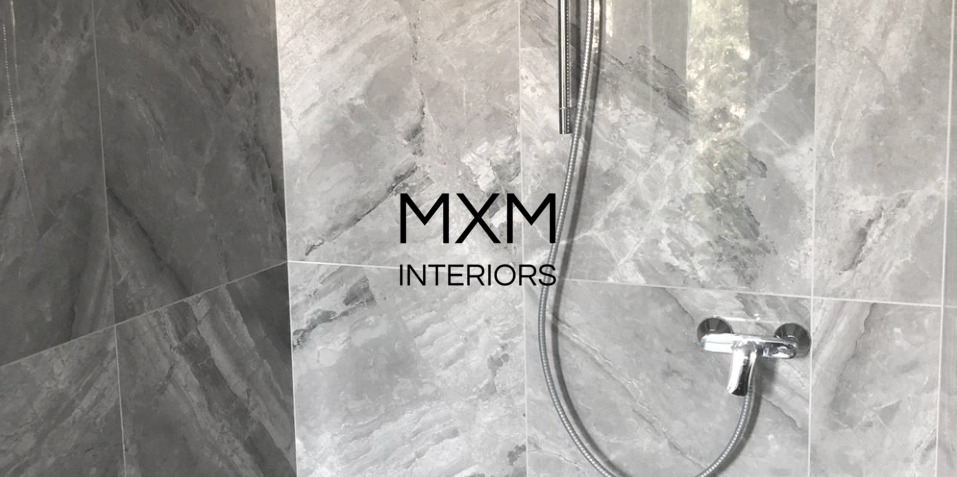 Szare marmurowe płytki w łazience z chromowaną baterią prysznicową i wężem; logo MXM Interiors nałożone cyfrowo.