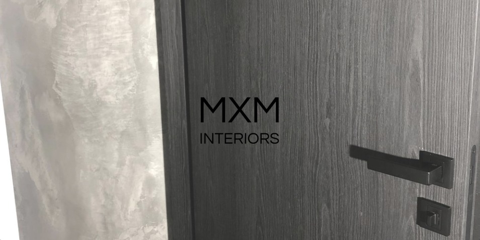 Ciemnoszare drzwi wewnętrzne z klamką i zamkiem w stylu minimalistycznym, na tle ściany z dekoracyjnym tynkiem imitującym marmur, z widocznym logo firmy MXM Interiors.