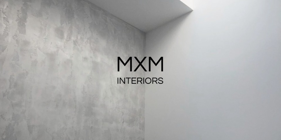 Ściana wykończona dekoracyjną szarą farbą strukturalną w rogu pomieszczenia, obok gładkiej, białej ściany, z widocznym logo firmy MXM Interiors.