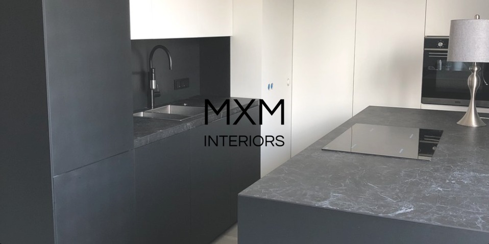 Nowoczesna kuchnia z ciemnoszarymi szafkami, czarną armaturą i marmurowym blatem, płyta indukcyjna w blacie wyspy kuchennej, oświetlenie punktowe, logo firmy MXM Interiors na pierwszym planie.