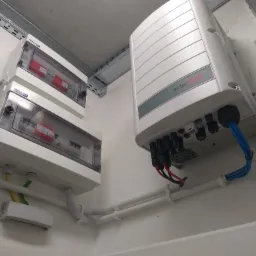 Instalacja fotowoltaiczna wewnątrz budynku: falownik SolarEdge z podłączonymi kablami, skrzynki zabezpieczeń z bezpiecznikami, kable w białych rurkach PCV.