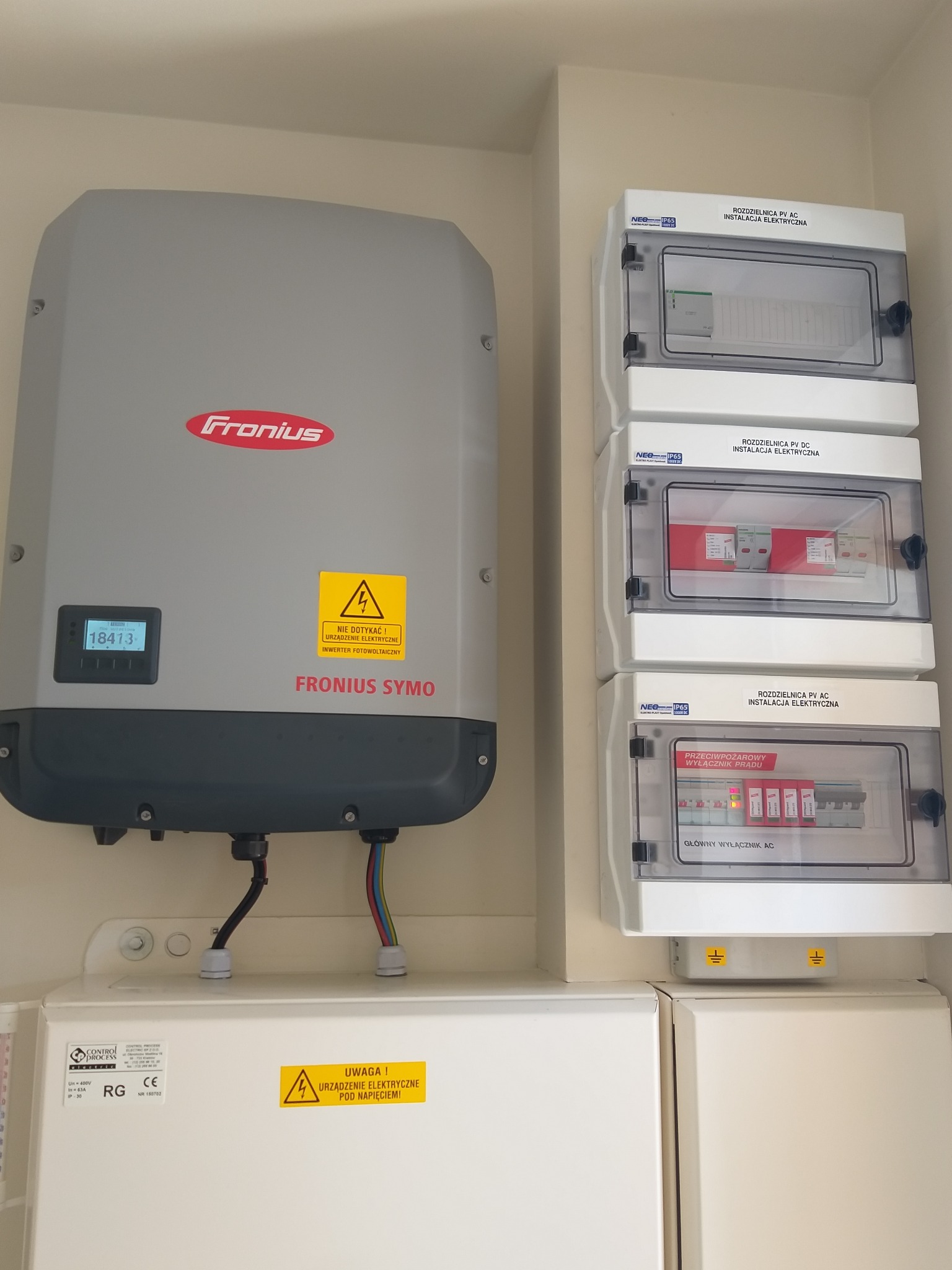 Szary inwerter Fronius SYMO z wyświetlaczem pokazującym produkcję energii, obok trzy białe rozdzielnice elektryczne PV AC i PV DC z przezroczystymi drzwiczkami, zamontowane na jasnej ścianie...