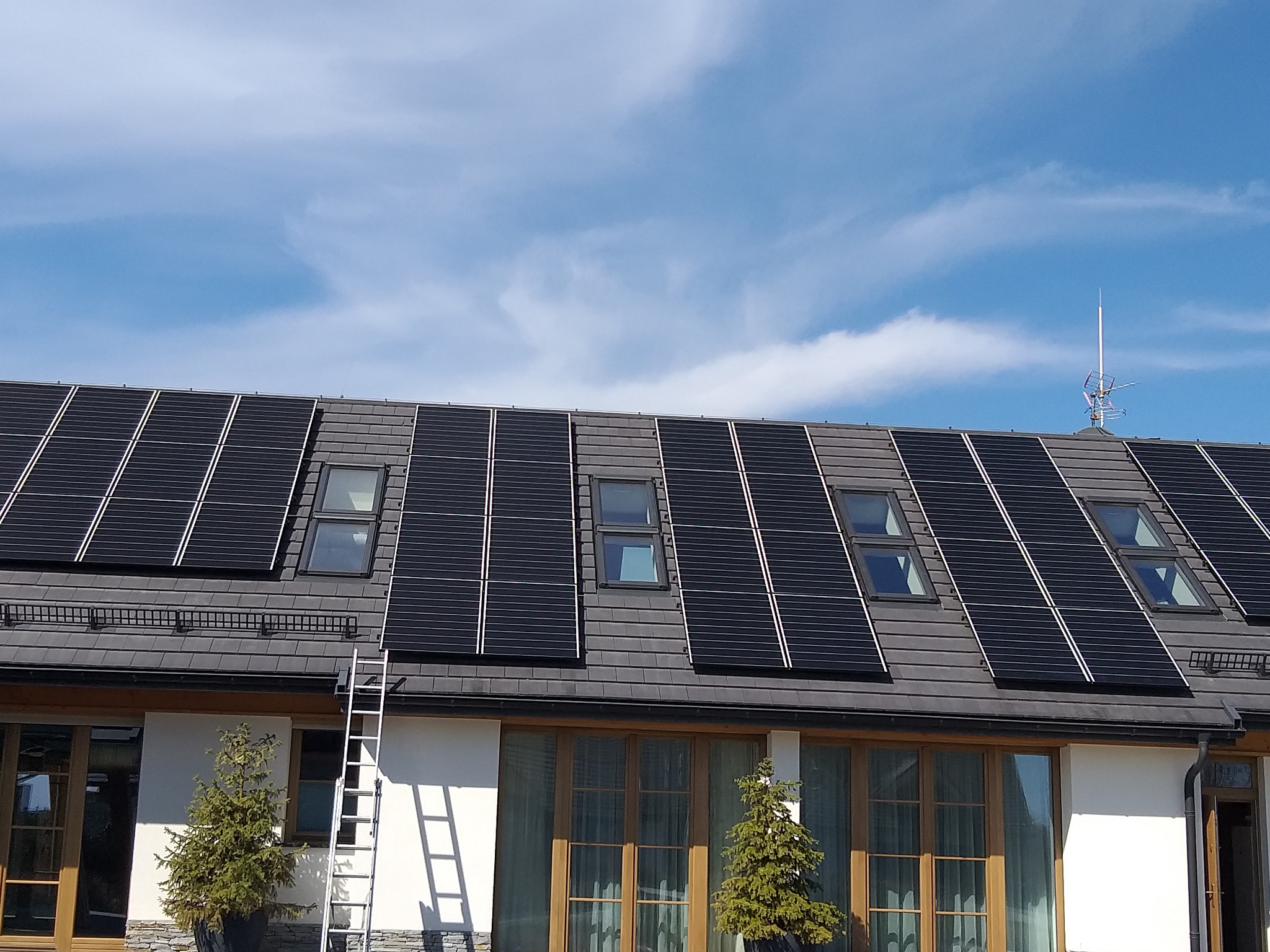 Czarny dach z panelami słonecznymi i oknami dachowymi, drabina oparta o ścianę budynku, w tle błękitne niebo.