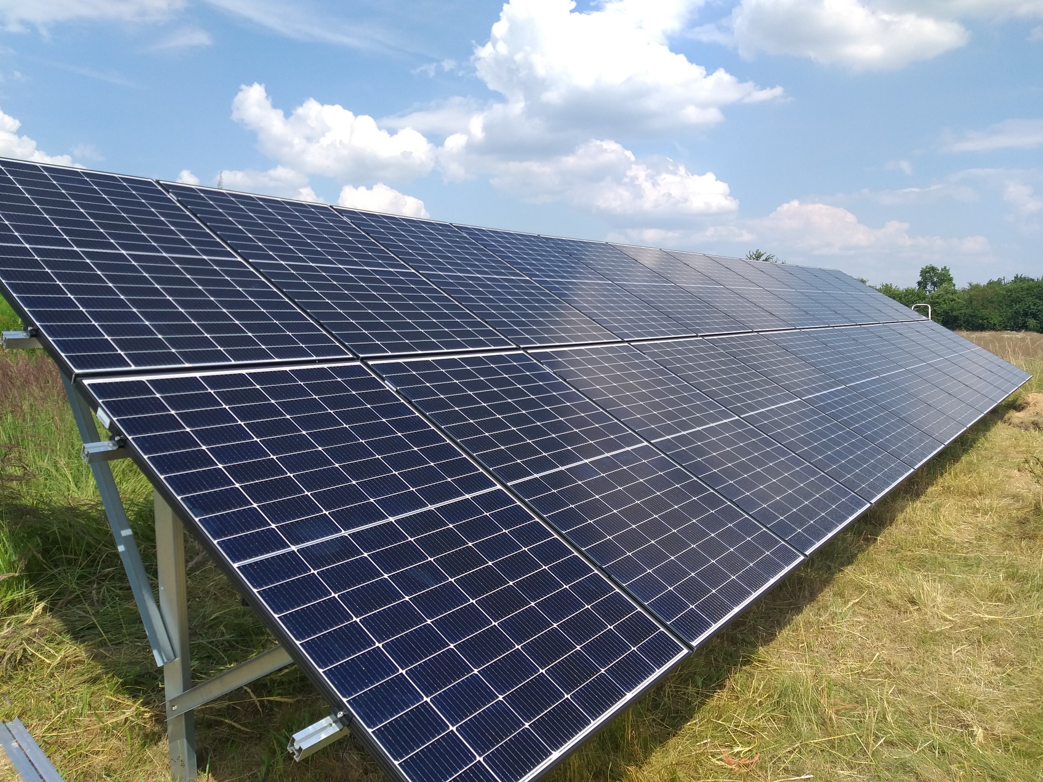 Rząd paneli słonecznych w trakcie instalacji na łące, widoczne metalowe podpory, błękitne niebo z chmurami w tle.
