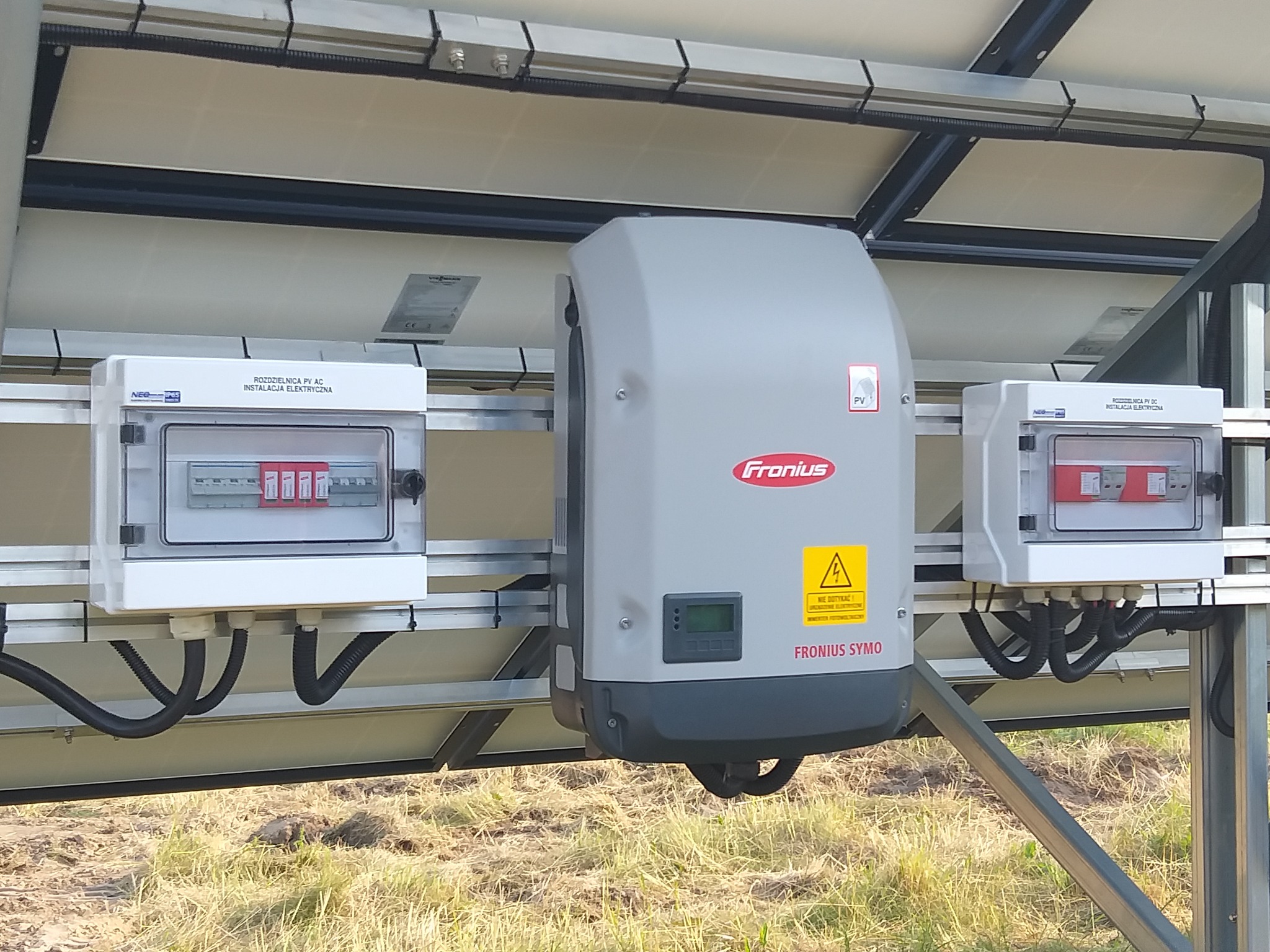 Instalacja fotowoltaiczna: Szary falownik Fronius SYMO z żółtą naklejką ostrzegawczą, dwie białe rozdzielnice AC/DC z widocznymi bezpiecznikami, zamontowane na konstrukcji paneli słonecznych na tle...
