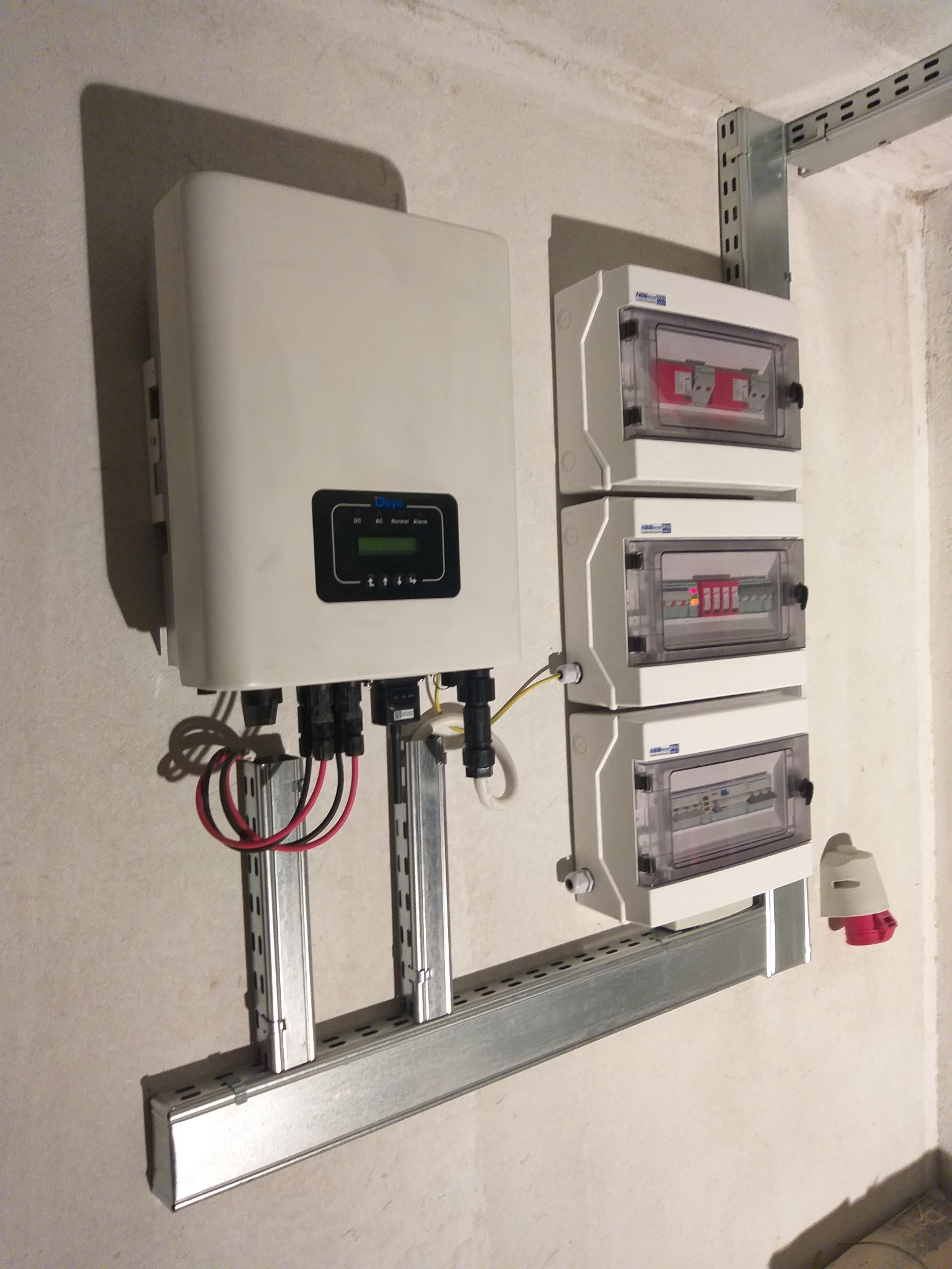 Biały inwerter fotowoltaiczny zamontowany na metalowej szynie, z widocznymi kablami i trzema skrzynkami rozdzielczymi z bezpiecznikami na ścianie o nierównej fakturze.