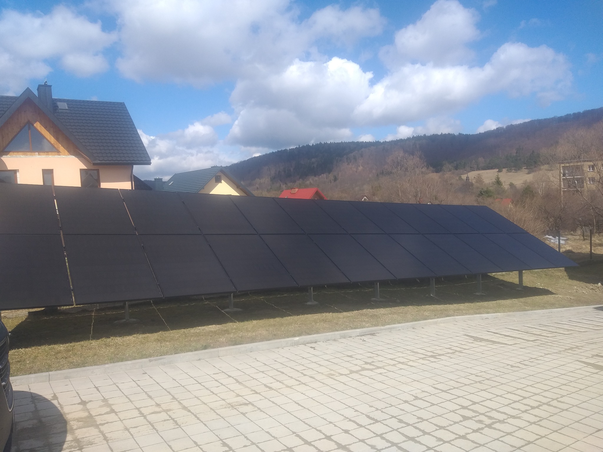 Długi rząd paneli fotowoltaicznych ustawionych na trawniku przed budynkami mieszkalnymi z widokiem na zalesione wzgórza w tle, widoczne ślady po deszczu na kostce brukowej.