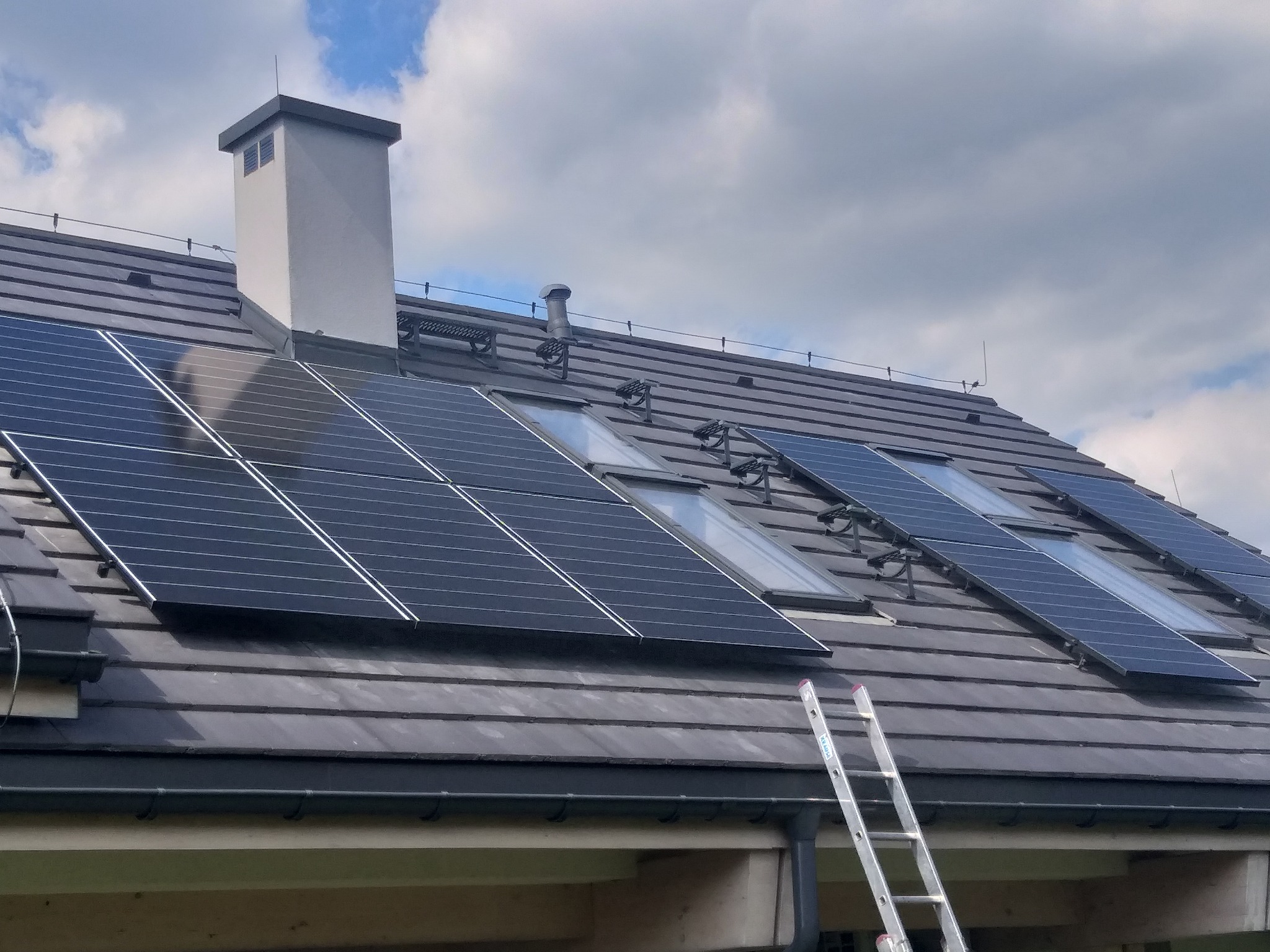 Częściowy widok dachu z dachówką ceramiczną, na którym zamontowano panele fotowoltaiczne i okna dachowe. Aluminiowa drabina oparta o rynnę.