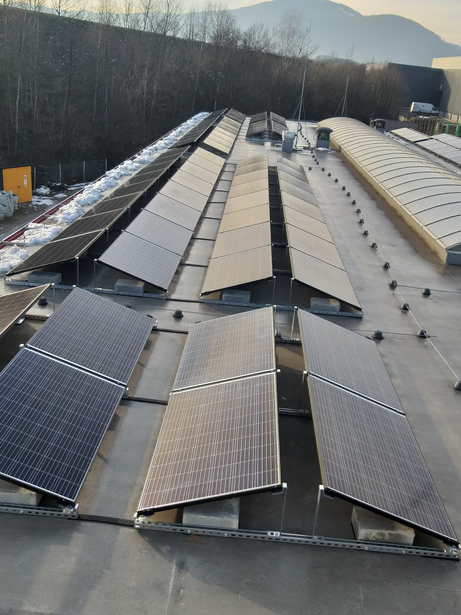 Rząd paneli słonecznych na płaskim dachu budynku przemysłowego, częściowo pokrytych śniegiem, z widocznymi mocowaniami i betonowymi podstawami, na tle zalesionych wzgórz i błękitnego nieba.