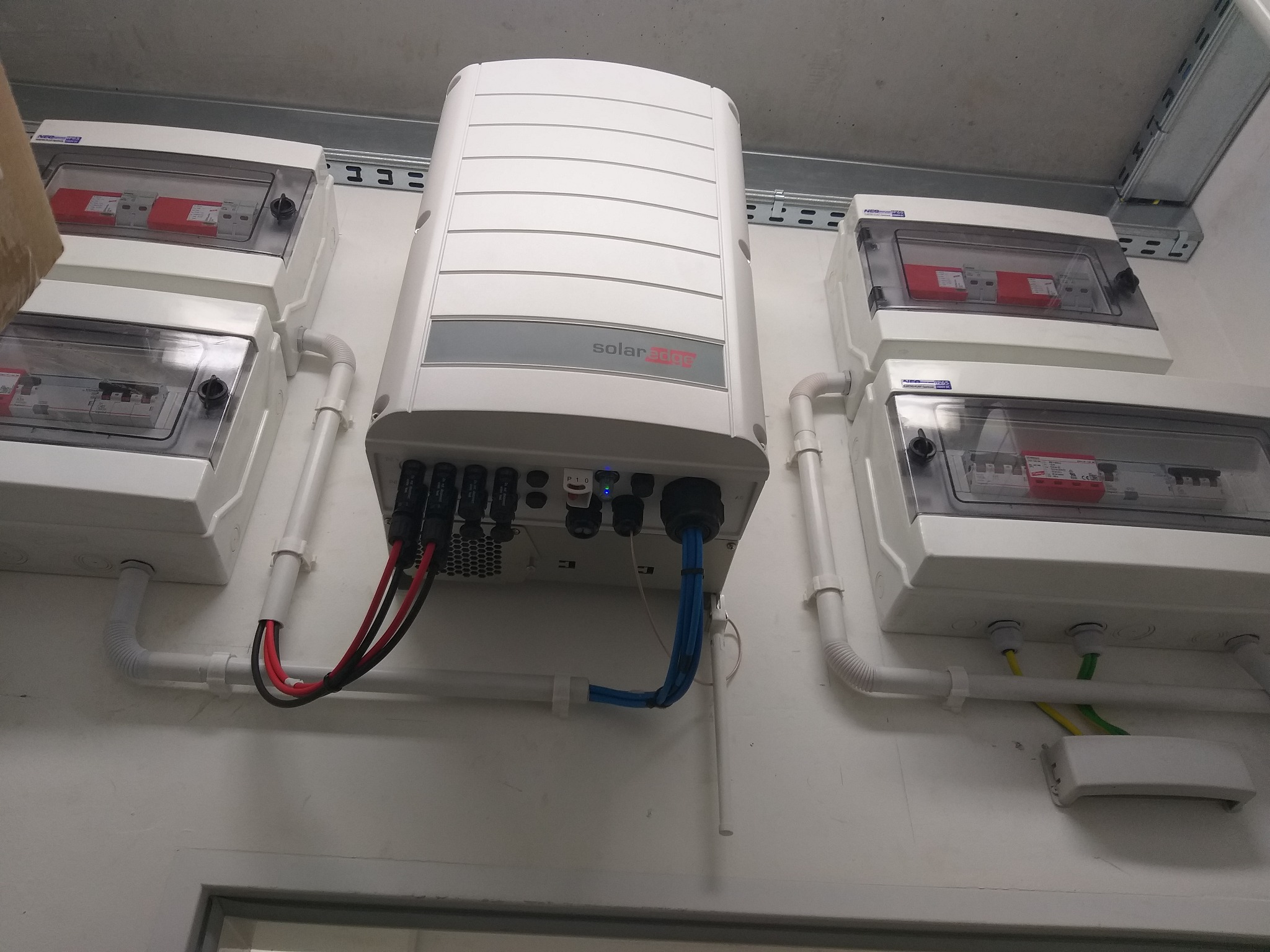 Biały inwerter SolarEdge zamontowany na ścianie obok czterech zabezpieczonych skrzynek elektrycznych, z widocznymi przewodami i rurami instalacyjnymi.