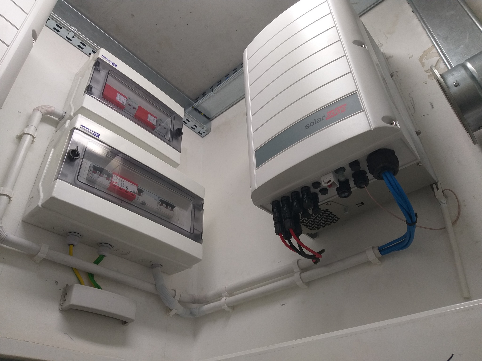 Instalacja fotowoltaiczna wewnątrz budynku: falownik SolarEdge z podłączonymi kablami, skrzynki zabezpieczeń z bezpiecznikami, kable w białych rurkach PCV.