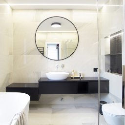 Hajder&Talarek - Nowoczesna łazienka z beżowymi płytkami imitującymi marmur, okrągłym lustrem z czarną ramą nad białą umywalką nablatową na czarnej szafce, kabiną prysznicową ze szklanymi drzwiami i białą wanną...