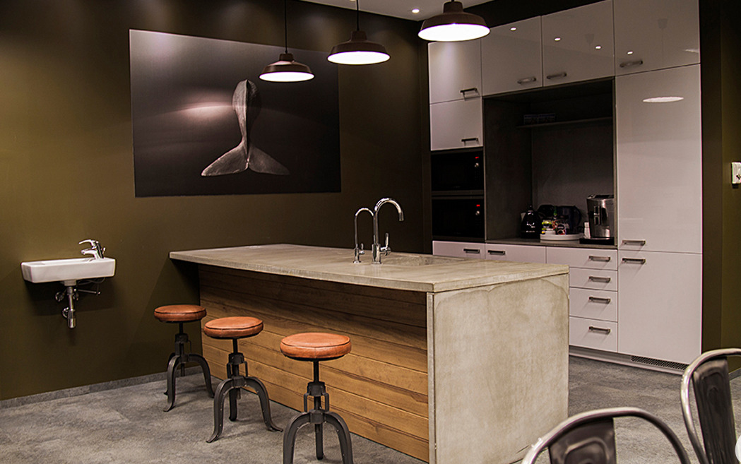 Wnętrze biura z wyspą kuchenną o betonowym blacie i drewnianym wykończeniu, trzema skórzanymi stołkami barowymi, białymi szafkami kuchennymi z wbudowanymi urządzeniami, umywalką na ścianie...