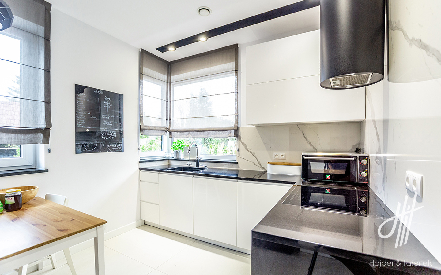 Jasna kuchnia w domu jednorodzinnym: białe szafki, czarny blat, okno z roletami rzymskimi, tablica kredowa z listą zakupów, nowoczesny okap cylindryczny.
