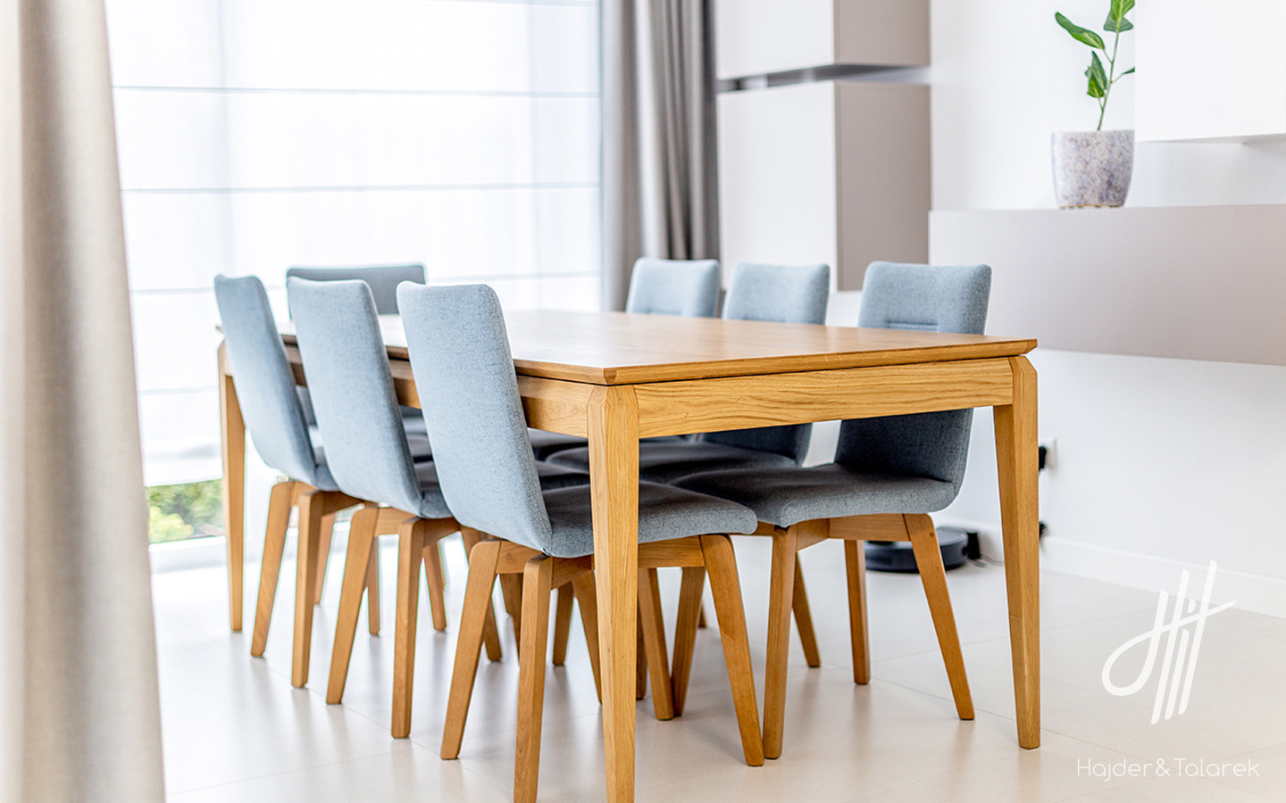 Stół z jasnego drewna i sześć krzeseł z szarobłękitną tapicerką w minimalistycznym wnętrzu domu jednorodzinnego, widok z boku, robot sprzątający w tle.