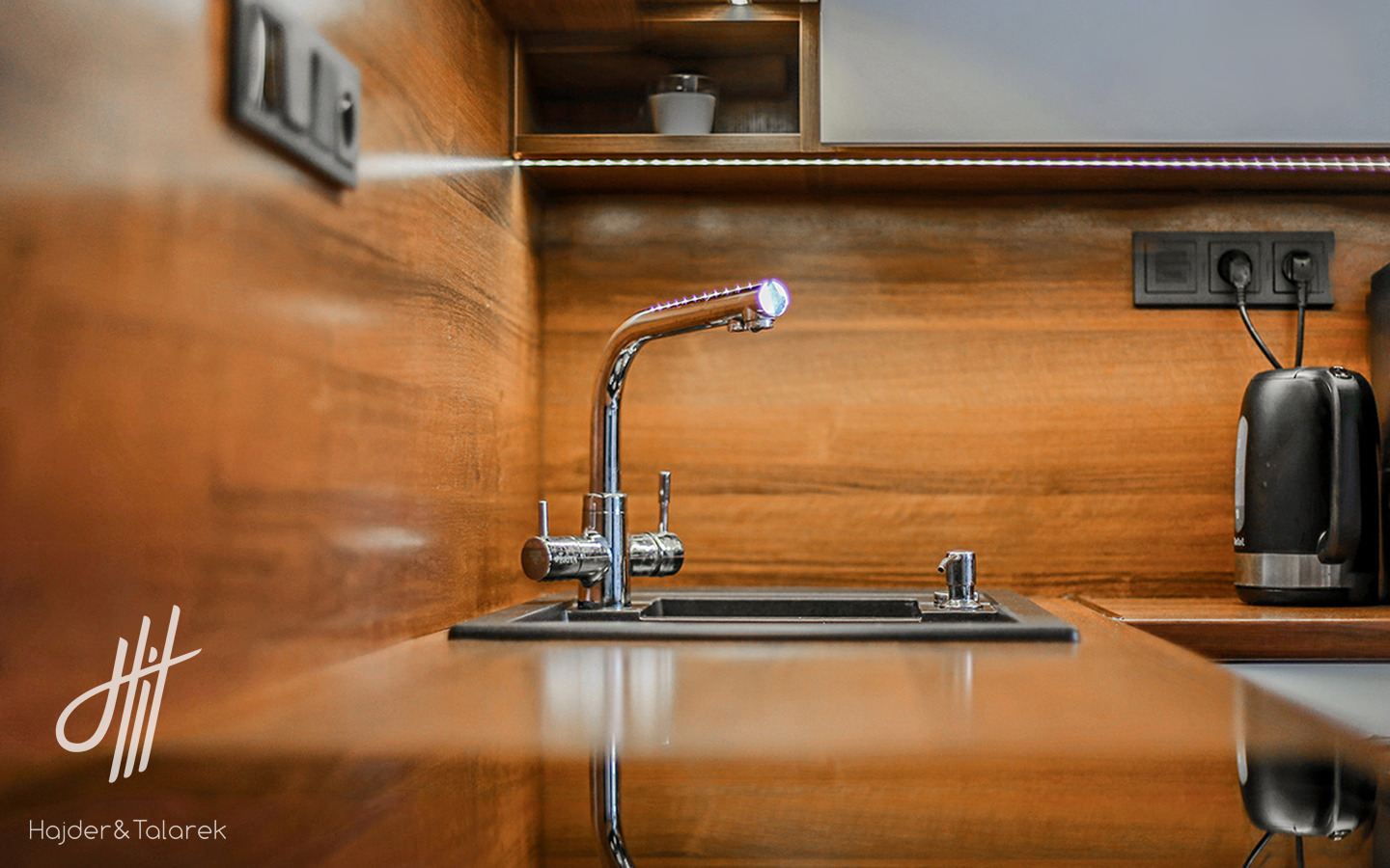 Nowoczesna bateria kuchenna z podświetleniem LED na tle drewnianej zabudowy z wbudowanym oświetleniem punktowym i czajnikiem elektrycznym. Widoczne gniazdka elektryczne i logo firmy projektowej...