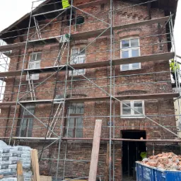 Renowacja ceglanej kamienicy z rusztowaniem, widoczni pracownicy w odblaskowych kamizelkach, worki z zaprawą murarską i kontener na gruz