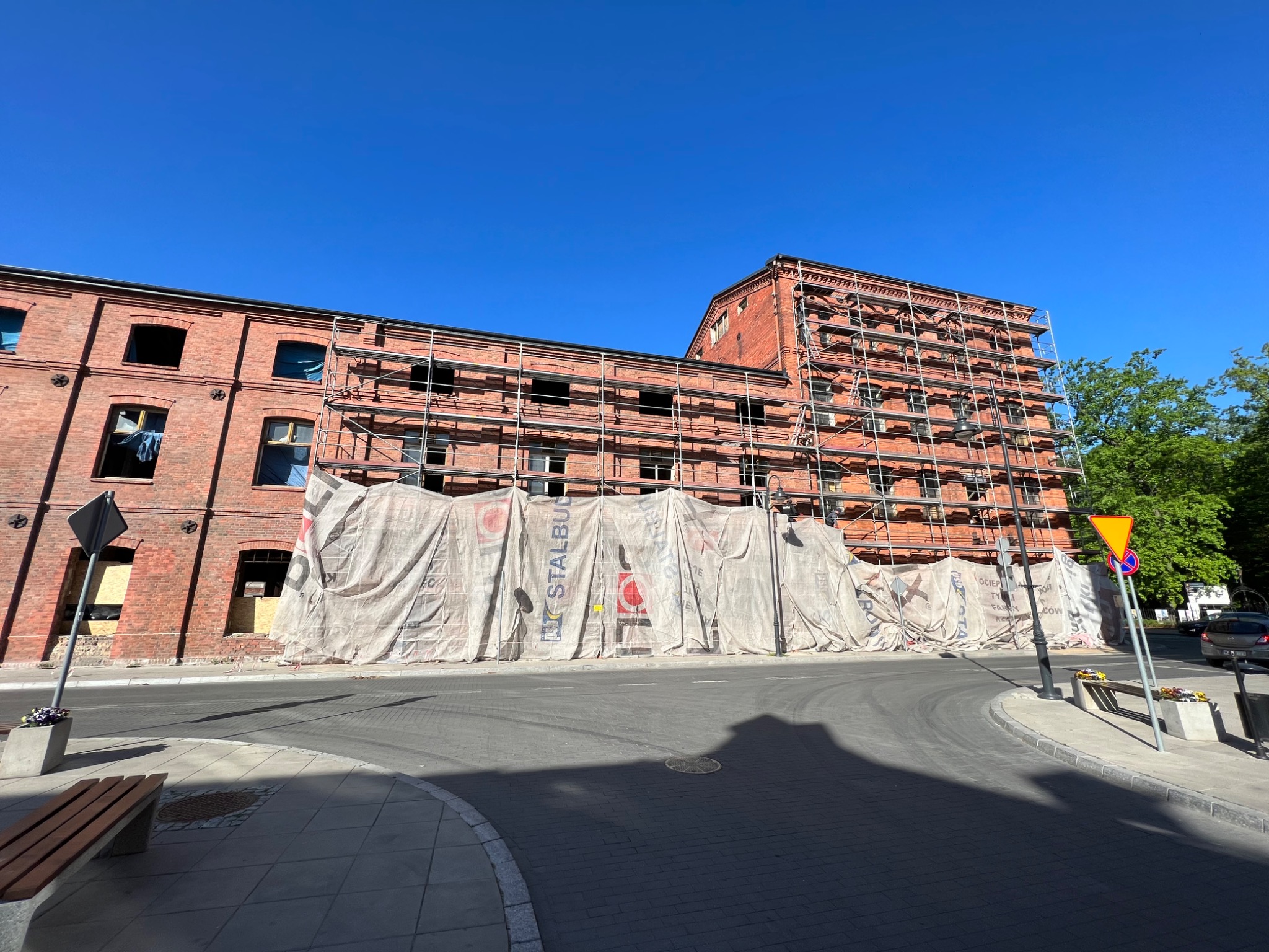 Ceglany budynek w trakcie renowacji, osłonięty plandekami z widocznymi rusztowaniami, ujęcie z poziomu ulicy w słoneczny dzień.