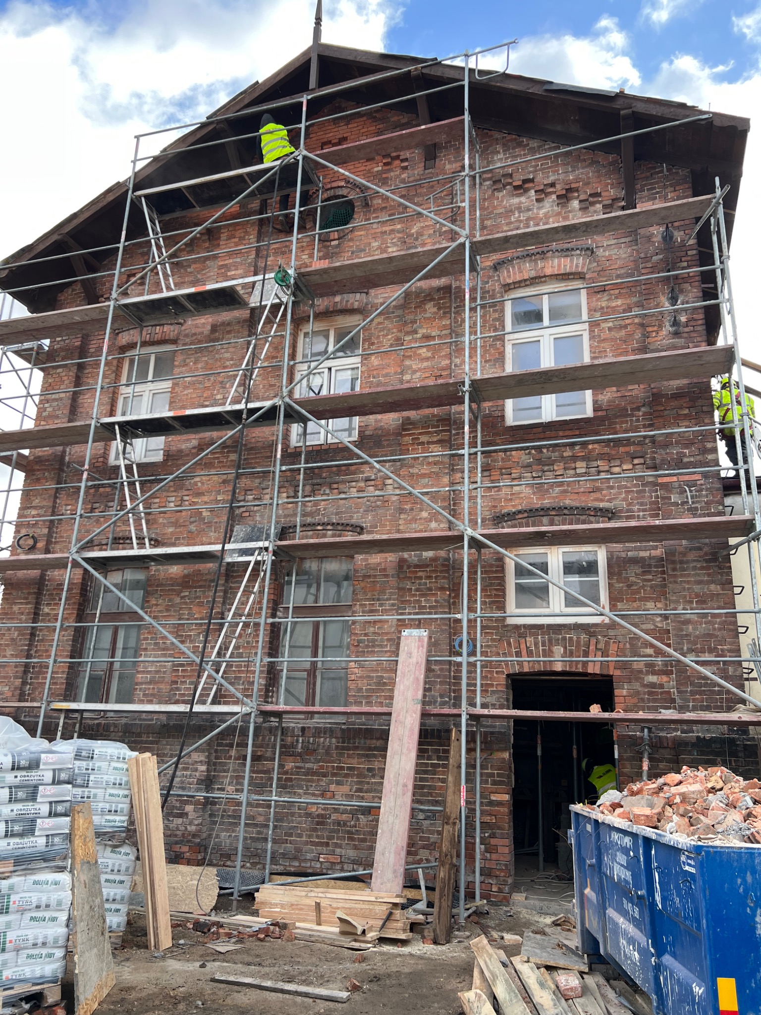 Renowacja ceglanej kamienicy z rusztowaniem, widoczni pracownicy w odblaskowych kamizelkach, worki z zaprawą murarską i kontener na gruz