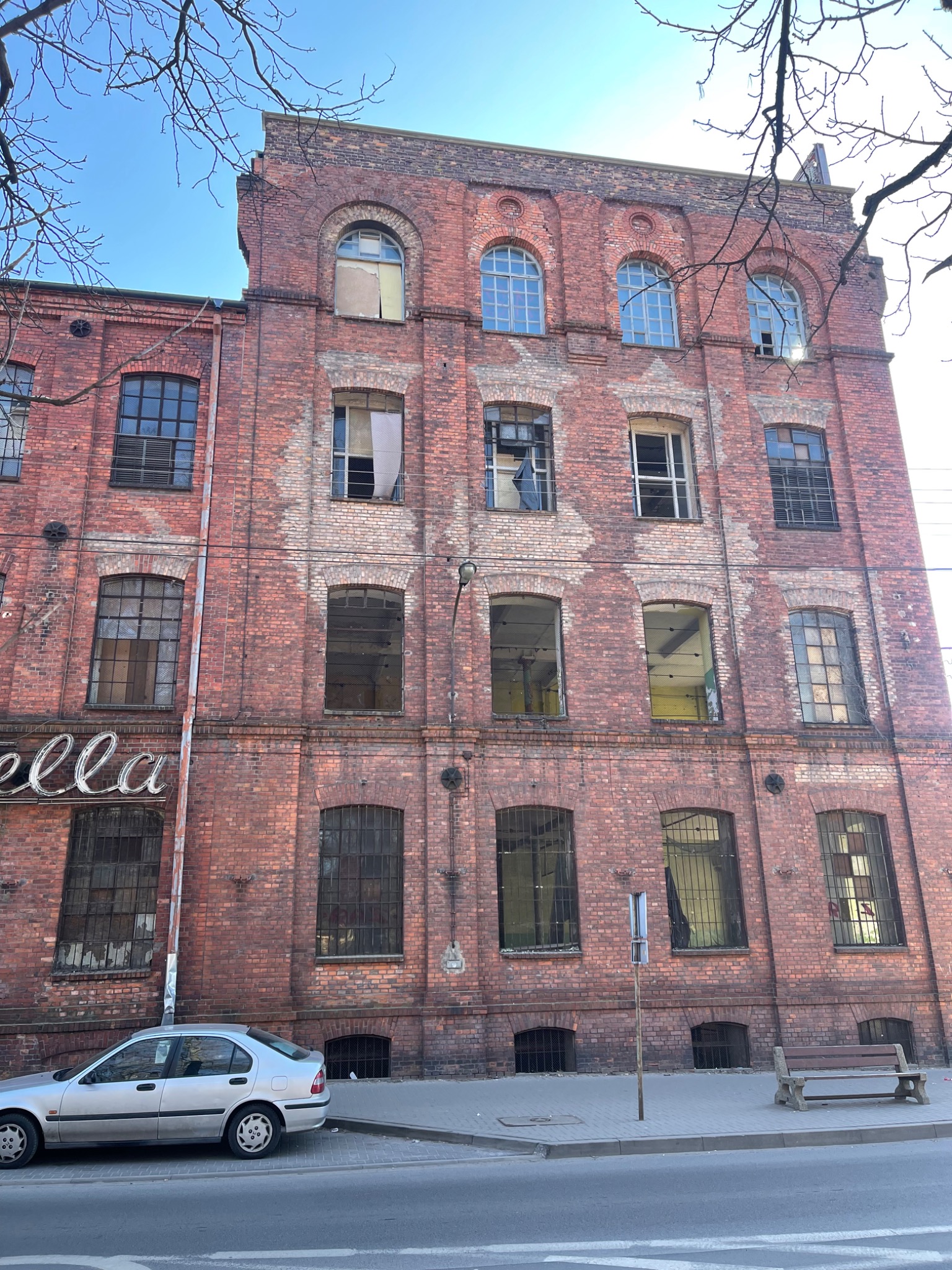 Wysoki, ceglany budynek z wieloma oknami, niektóre z nich są uszkodzone lub zabite deskami, z widocznym napisem 'ella' na fasadzie, zaparkowany samochód i ławka na chodniku.