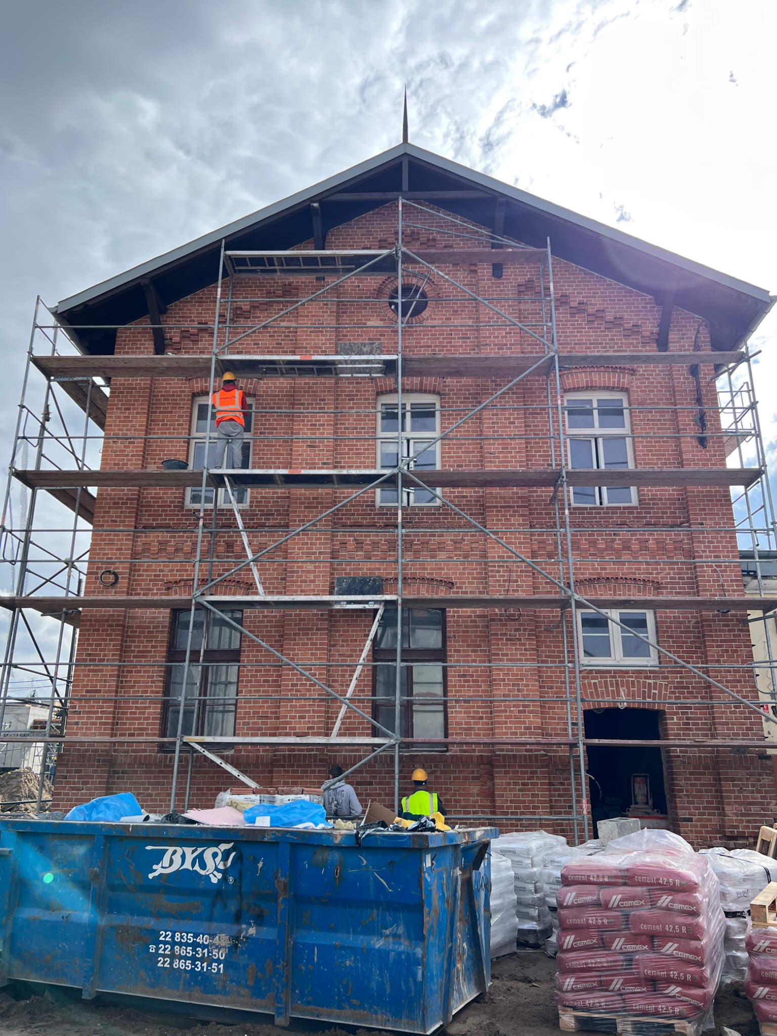 Renowacja zabytkowej, ceglanej kamienicy z rusztowaniami, widoczny pracownik w kamizelce odblaskowej na rusztowaniu, kontener na gruz i palety z cementem na pierwszym planie.