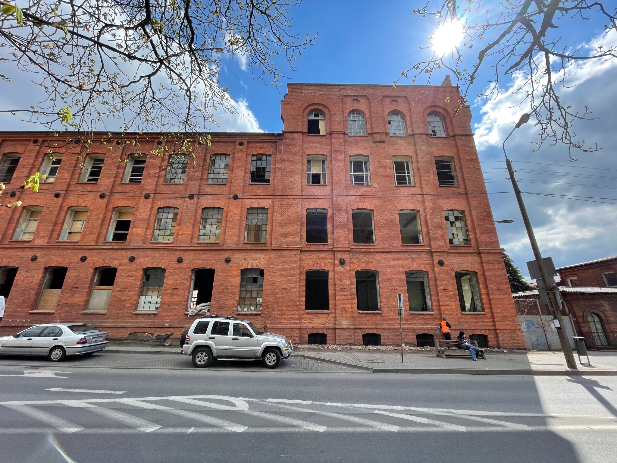 Ceglany budynek fabryczny z powybijanymi oknami, widok z ulicy z zaparkowanymi samochodami i osobą siedzącą na ławce w pomarańczowej kamizelce odblaskowej, fragment nieba z promieniami słońca...