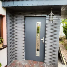 Szare drzwi wejściowe z wąskim, pionowym przeszkleniem i chromowaną klamką, umieszczone w wejściu wyłożonym cegłopodobnymi płytkami, zwieńczonym daszkiem z dachówki.