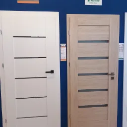 Dwie nowoczesne drzwi wewnętrzne: białe z czarną klamką i jasne drewnopodobne ze srebrną klamką, prezentowane na tle niebieskiej ściany w salonie z logo producentów.