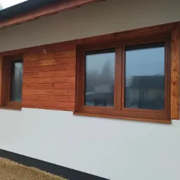 Dwa okna z drewnianą ramą w nowym budynku z jasną elewacją i drewnianą boazerią pomiędzy nimi. Widok na dom sąsiada w odbiciu szyb.