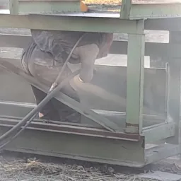 Pracownik w kombinezonie ochronnym, z bliska, podczas czyszczenia metalowej konstrukcji strumieniem ścierniwa, widoczne przewody i pył.