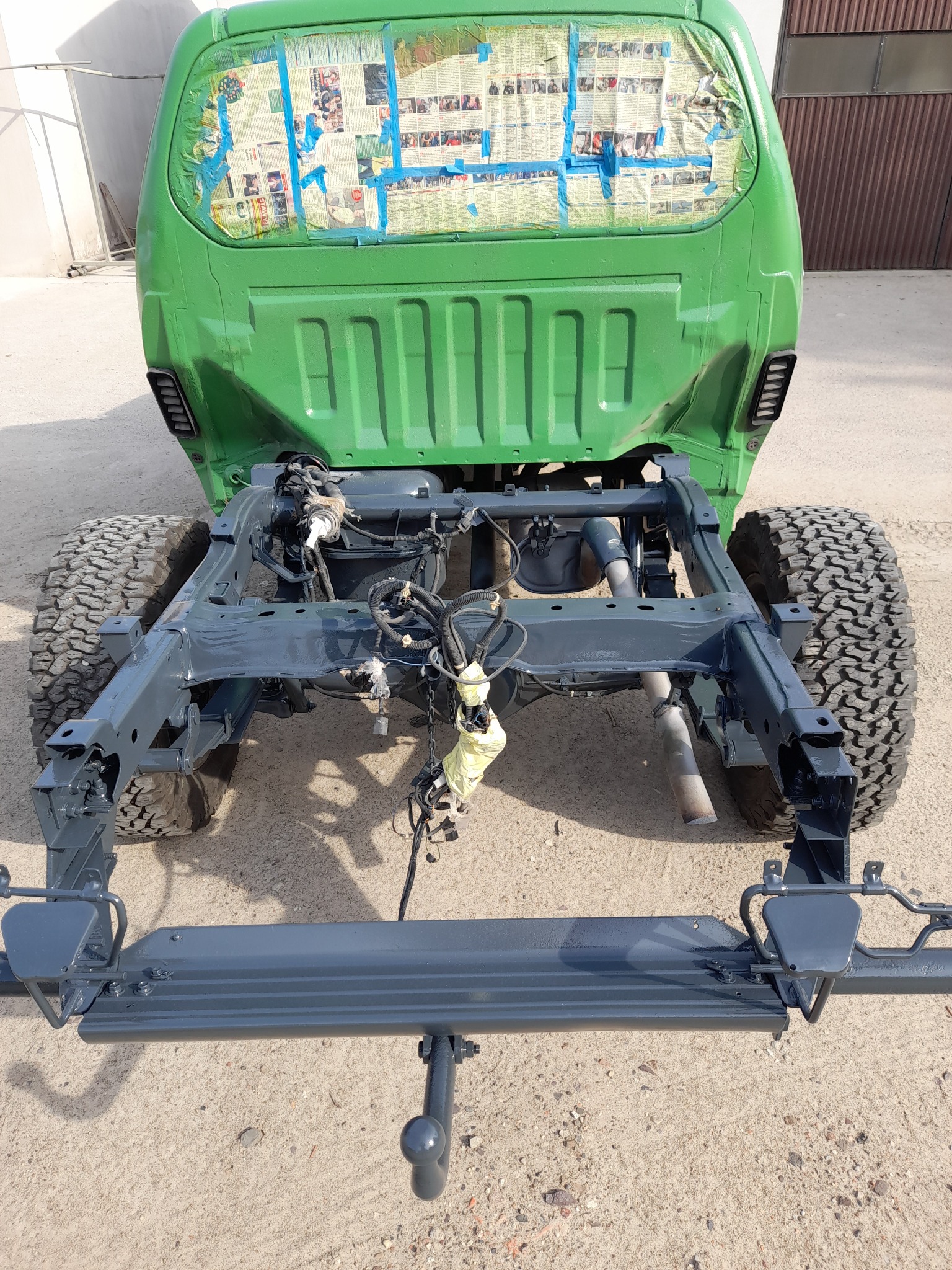 Tylny widok podwozia samochodu terenowego po renowacji, z elementami pomalowanymi na szaro i kabiną w odcieniu zieleni, zabezpieczoną gazetami przed malowaniem.