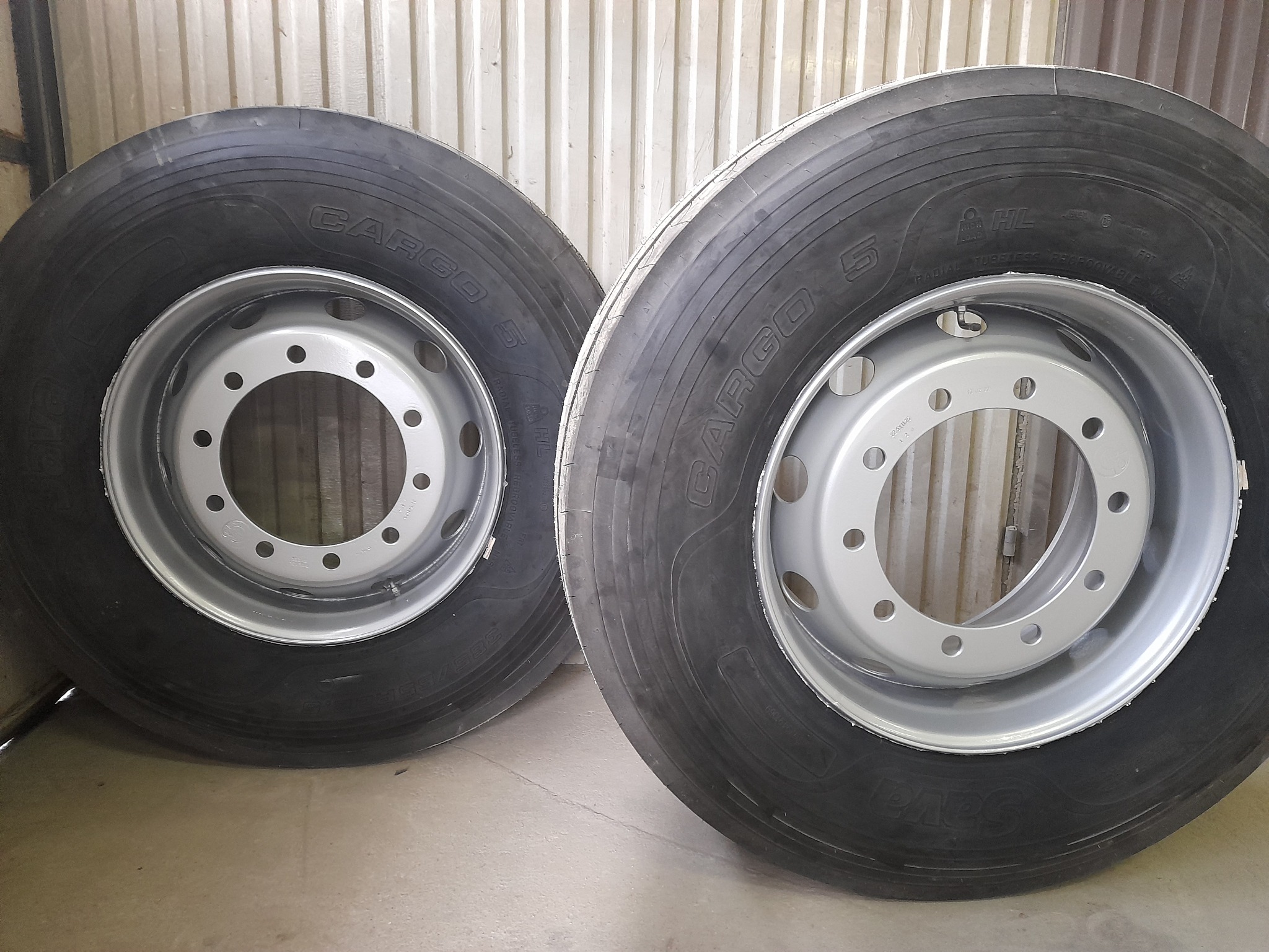 Dwa koła ciężarowe z oponami, felgi w kolorze srebrnym, opony czarne z widocznymi oznaczeniami producenta i modelu, ustawione na betonowej podłodze przed metalową ścianą z blachy falistej.