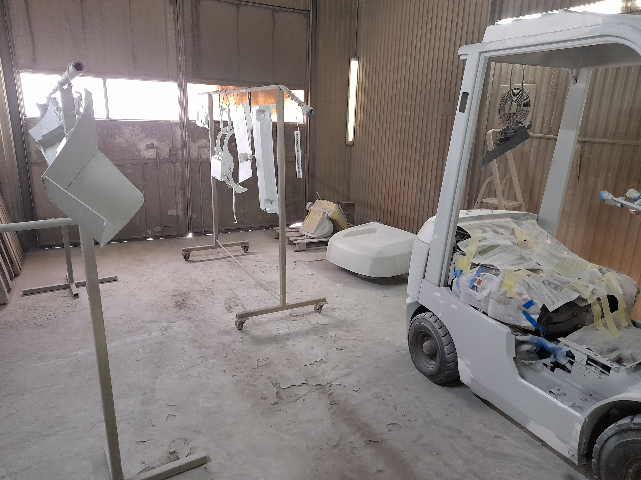 Wnętrze warsztatu z elementami po piaskowaniu i malowaniu, w tym wózek widłowy częściowo zabezpieczony papierem, metalowe konstrukcje z zawieszonymi detalami oraz inne elementy pojazdów...
