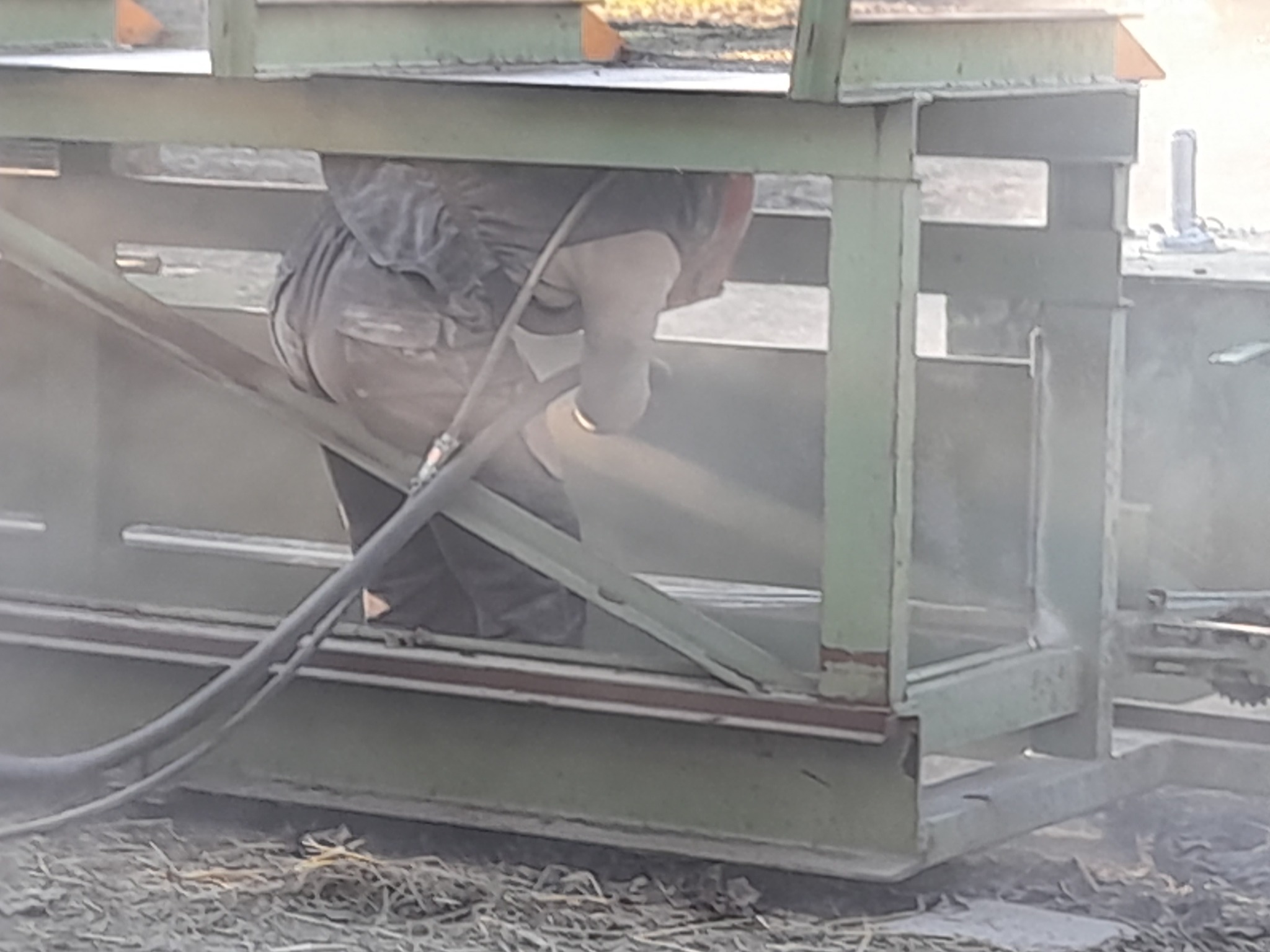 Pracownik w kombinezonie ochronnym, z bliska, podczas czyszczenia metalowej konstrukcji strumieniem ścierniwa, widoczne przewody i pył.