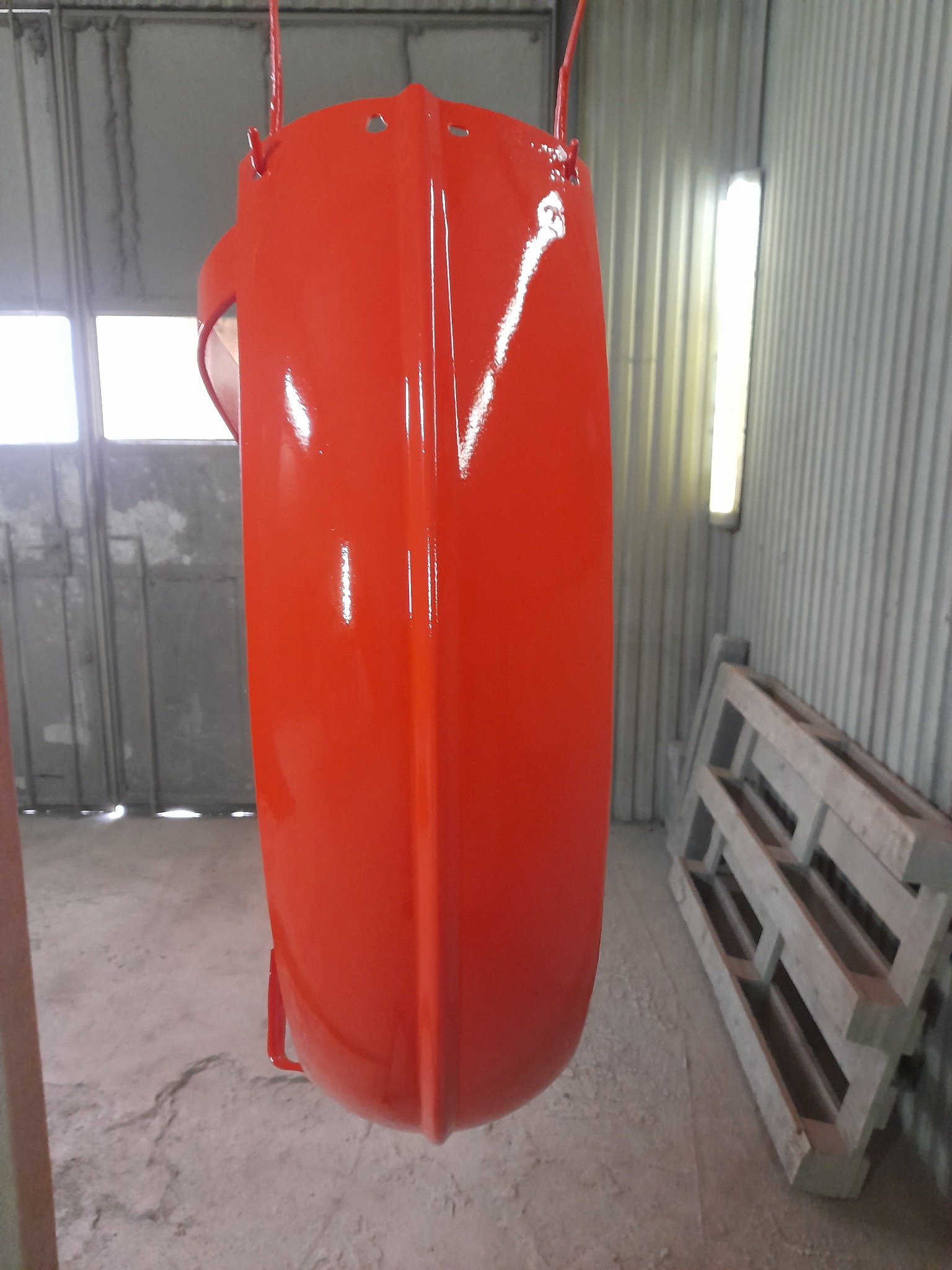 Czerwony błotnik motocyklowy świeżo po malowaniu proszkowym, zawieszony na lince w warsztacie z widocznymi paletami w tle.